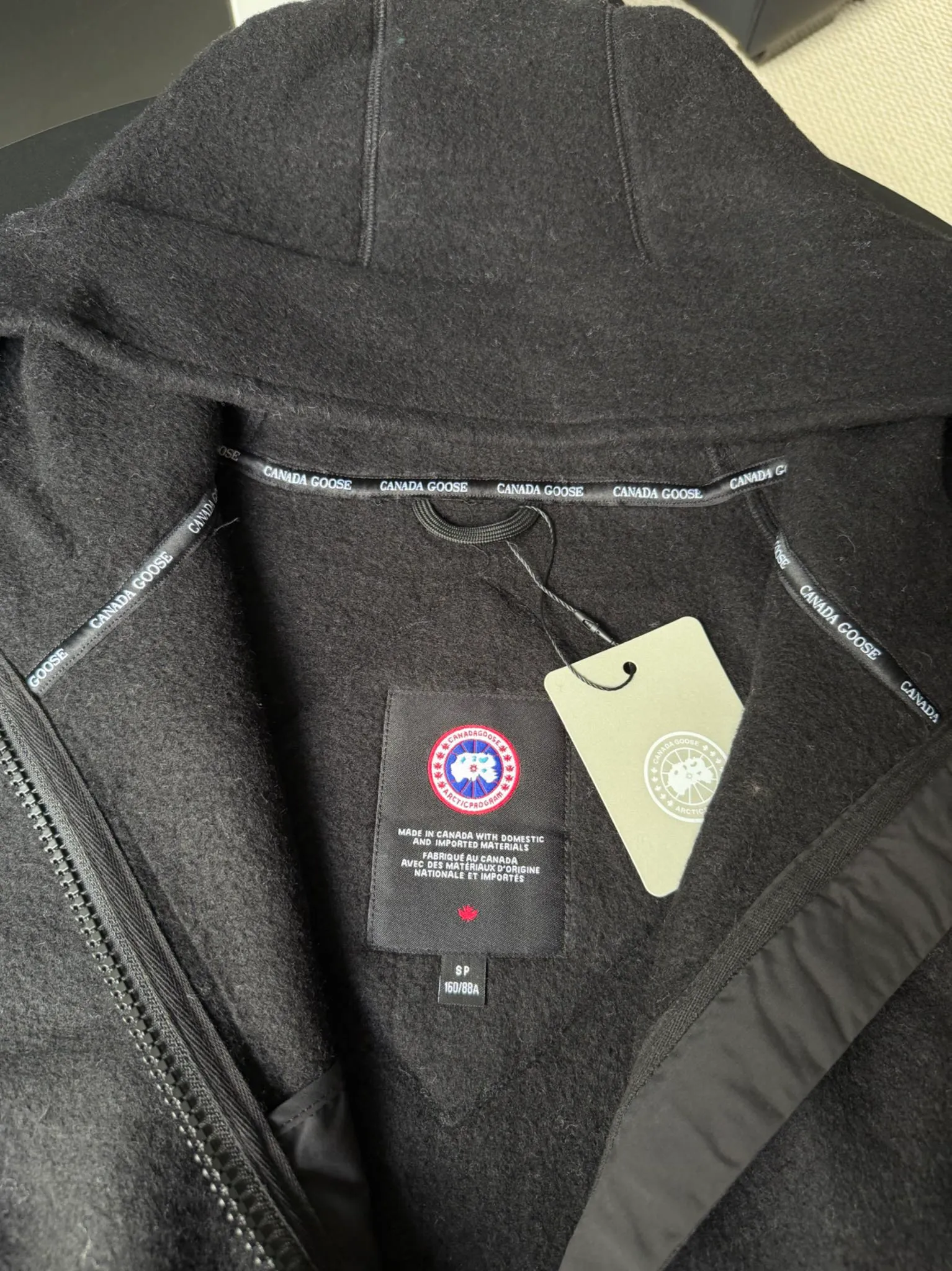 P660 Canada Goose Fleece Wool Badge Hooded Jacket Fabric selection: Wool fleece fabric blended with wool as the main body and a very small amount of spandex and polyester fiber. Wool is a core natural ingredient that provides unparalleled temperature regulation, antibacterial and anti-odor properties and a skin-friendly feel. The feeding of a small amount of spandex gives the fabric a certain degree of four-way elasticity, making it non-restrictive during daily activities. As the basis of fleece's structure, polyester fiber enhances the fabric's durability, wrinkle resistance and quick-drying properties. It contributes to the loft of the fleece and provides functions such as trapping air.  Detail 1: The sleeves and hem are closed with 2×2 ribbing. Its strong elasticity can closely fit wrists and waists of different thicknesses, forming an effective 'seal' to prevent cold wind from blowing back. It can still maintain its original shape after repeated stretching and use, and is not easy to loose and deform. And the ribbed cuffs can convey a sense of design and enhance the visual beauty.  Detail 2: The combination of hood design and elastic buckle improves warmth and windproof performance when wearing. Accessories: Customized elastic drawstring, elastic buckle, custom zipper pendant, custom armband Size: S M L Color: black gray coffee rice pink